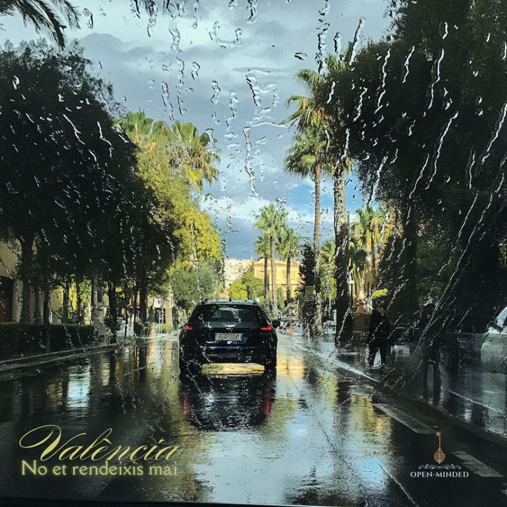 Miércoles 30 Octubre Cerrados. No et rendeixis mai, València! 1 Vista de una calle en Valencia bajo la lluvia, con gotas de agua en el parabrisas que distorsionan la imagen de palmeras y un cielo nublado. Texto en la esquina inferior izquierda: ‘València, No et rendeixis mai.’ Logo de OPEN-MINDED en la esquina inferior derecha
